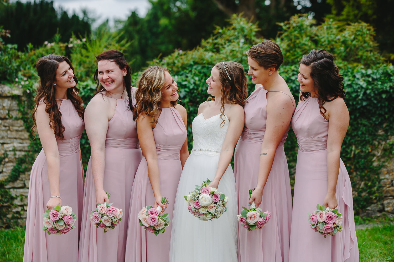 A bride and her bridesmaid smiling at each other while standing in a row.