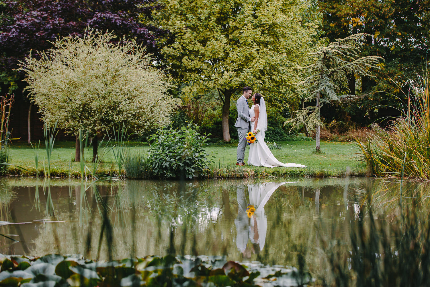 Bride and groom by a lake, reflection in water, grass, trees