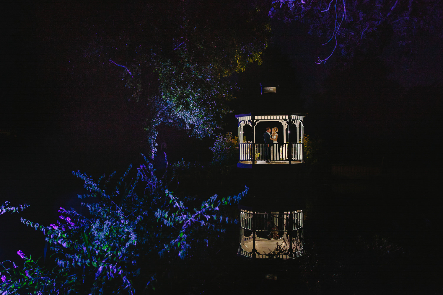 Bride and groom, night time, gazebo, purple lit trees, lake, reflection