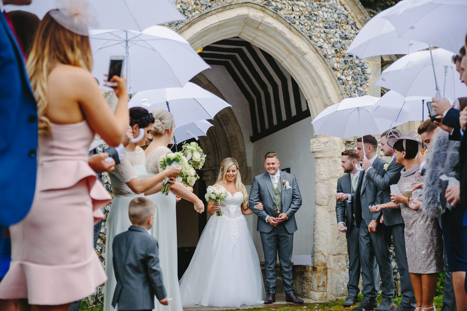 Bride and groom in church arch, weddings guests with umbrellas