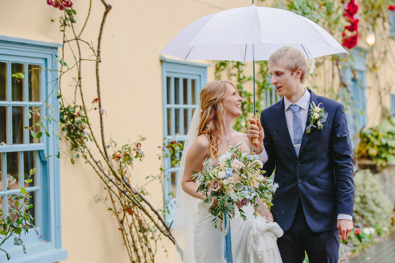Bride and groom, white umbrella in front of yellow house with blue windows