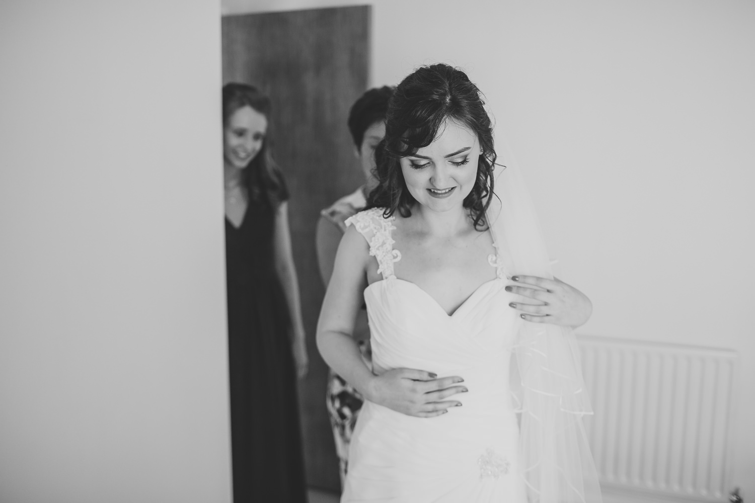 Black and white wedding photo of bride getting into her wedding dress being helped by her mum and bridesmaids