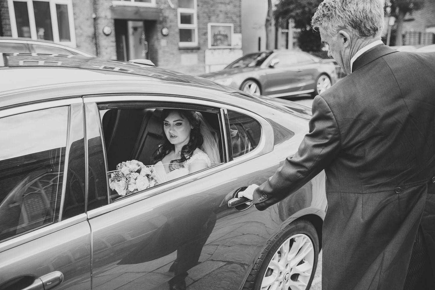 Black and white photo of a bride arriving by car to her wedding day with the door being opened by her father
