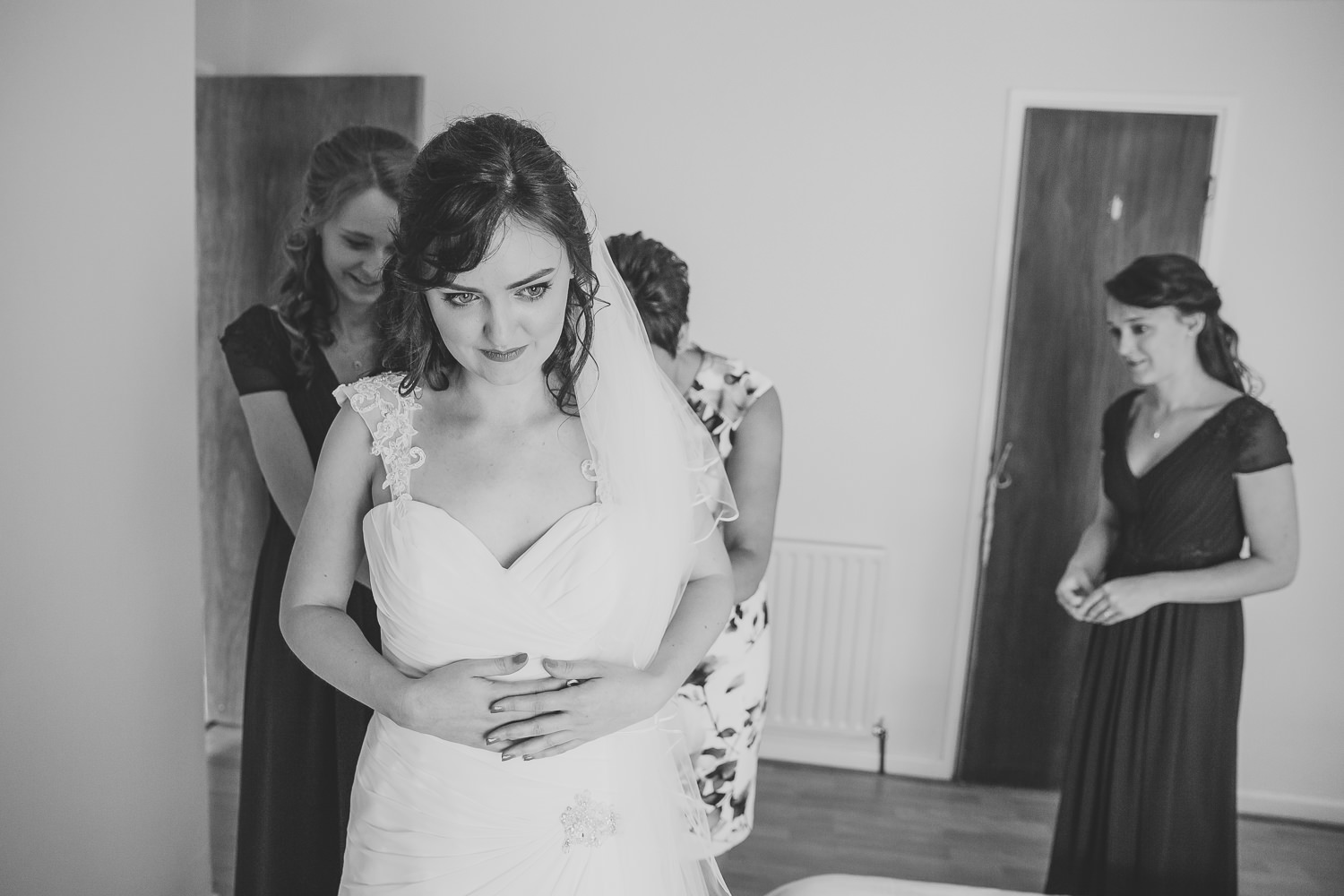 Black and white wedding photo of bride getting into her wedding dress being helped by her mum and bridesmaid