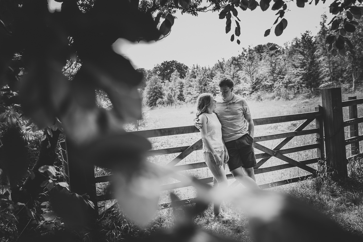 Black and white photo of an engaged couple leaning against a gate in Wandlebury Country Park in Cambridge.