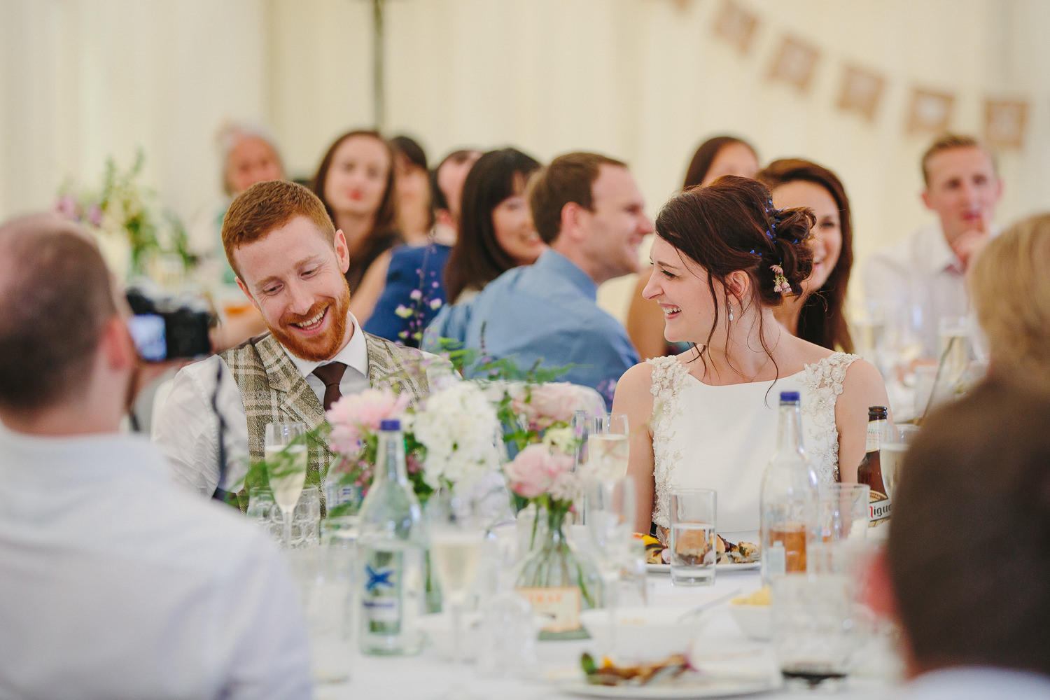 Bride and groom looking at each other smiling during wedding speeches, inside white marquee. Photographed by an Ely Wedding Photographer