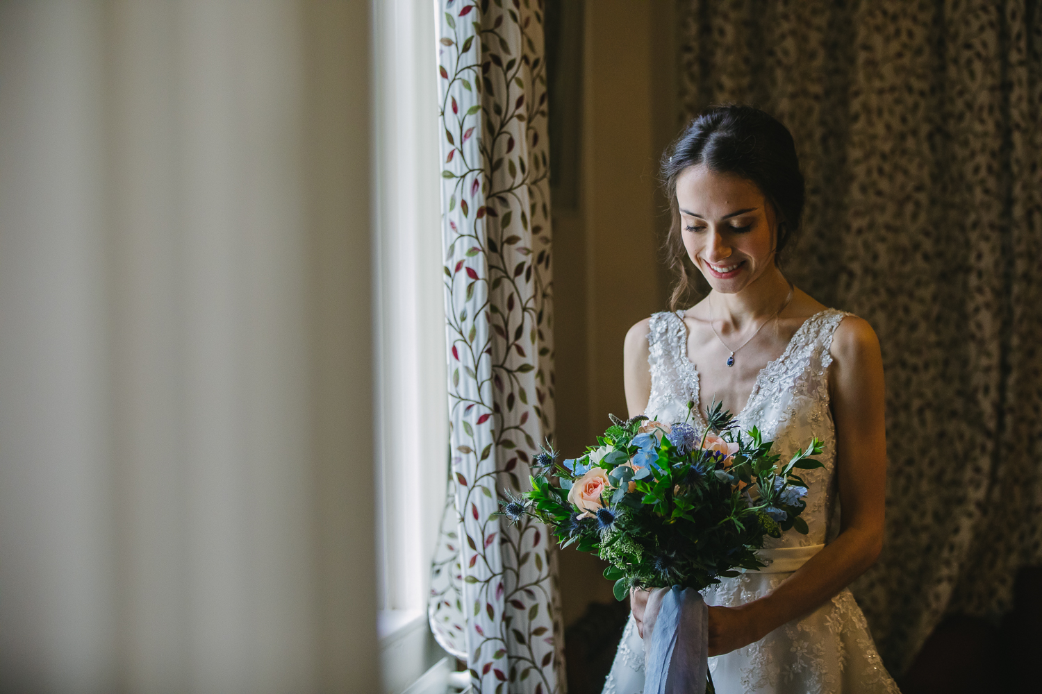 Bridal portrait, bride standing by a window looking at her bouquet in side Magdalene college Cambridge. Photo by Cambridge University Wedding Photographer