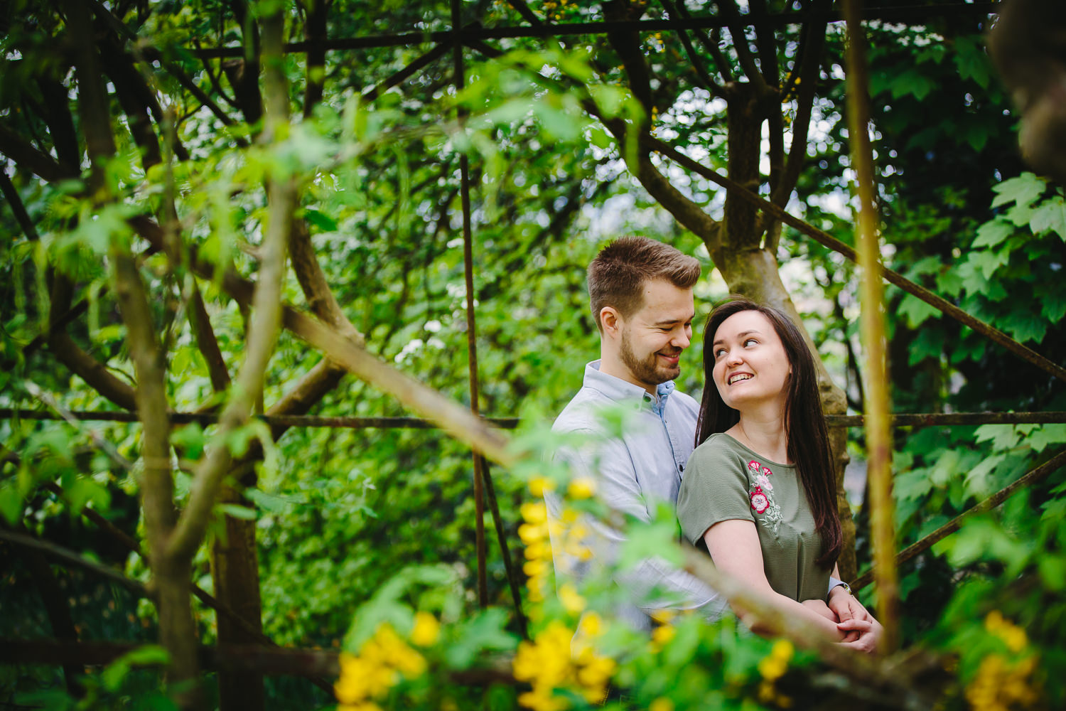 Richmond engagement shoot photo of a couple smiling at one another, under an archway covered in foliage and yellow flowers