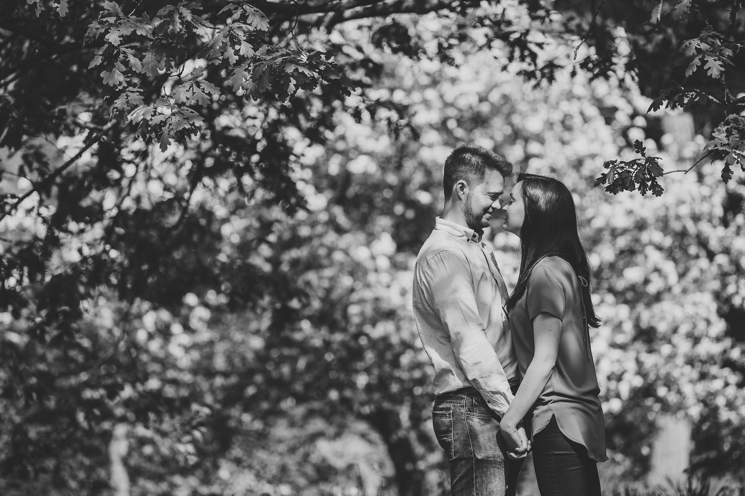 Black and white Richmond engagement shoot photo of a couple smiling at one another, amongst the trees in the woodland of Richmond Park.