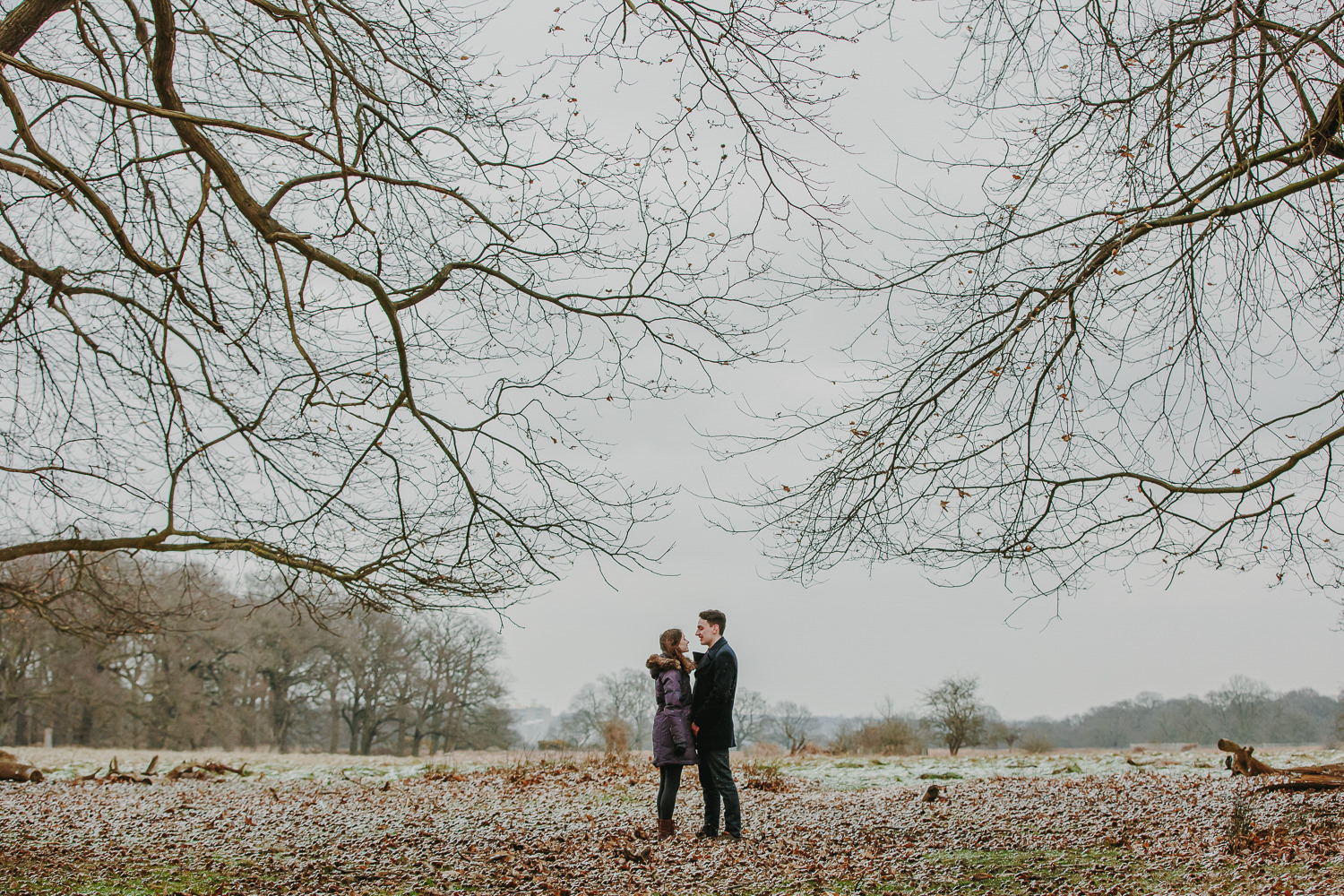 Engaged couple standing underneath trees at Richmond Park London. The ground is covered in snow.