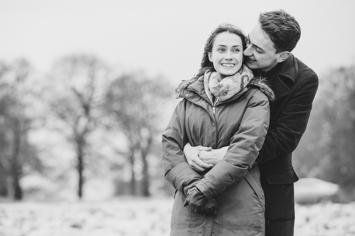 Black and white engagement photo of couple standing together in Richmond Park, which is covered in snow.
