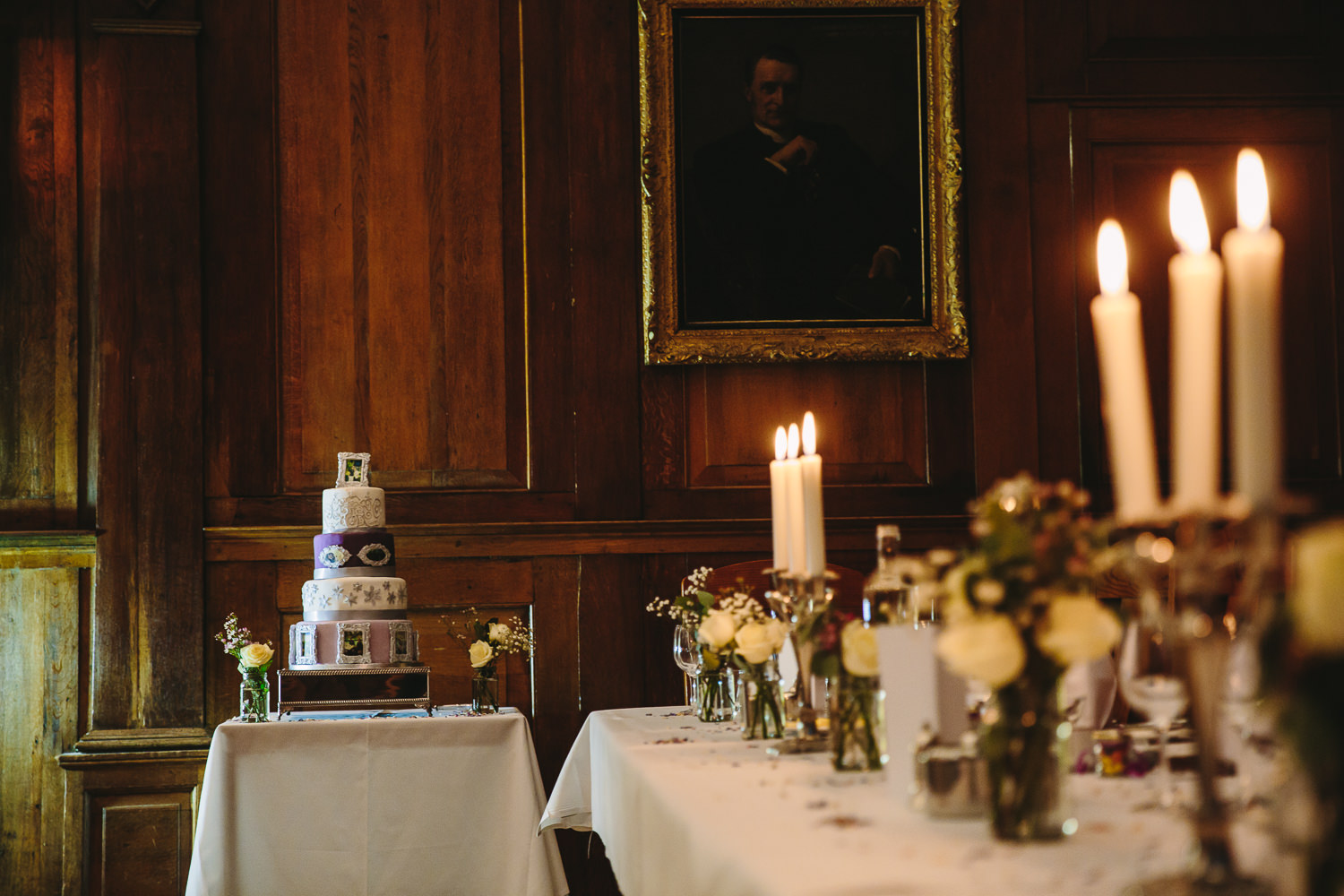 Purple and white Wedding cake situated next to a table decorated with flowers and candles inside Cambridge University Selwyn College