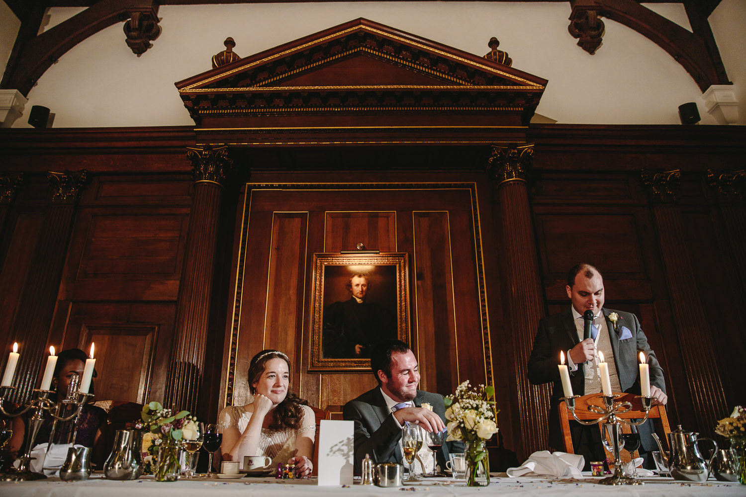 Wedding speeches inside the main hall at Selwyn college Cambridge university. Bride, groom and bridesmaid listen to the best man speech with animated expressions.