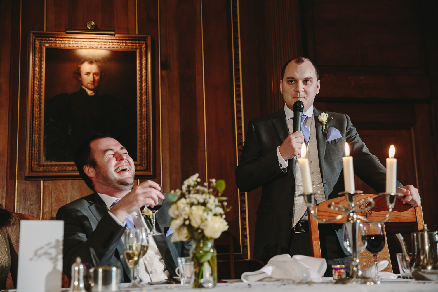 Best man giving a wedding speech inside the candlelit hall at selwyn college Cambridge university. Groom is laughing.