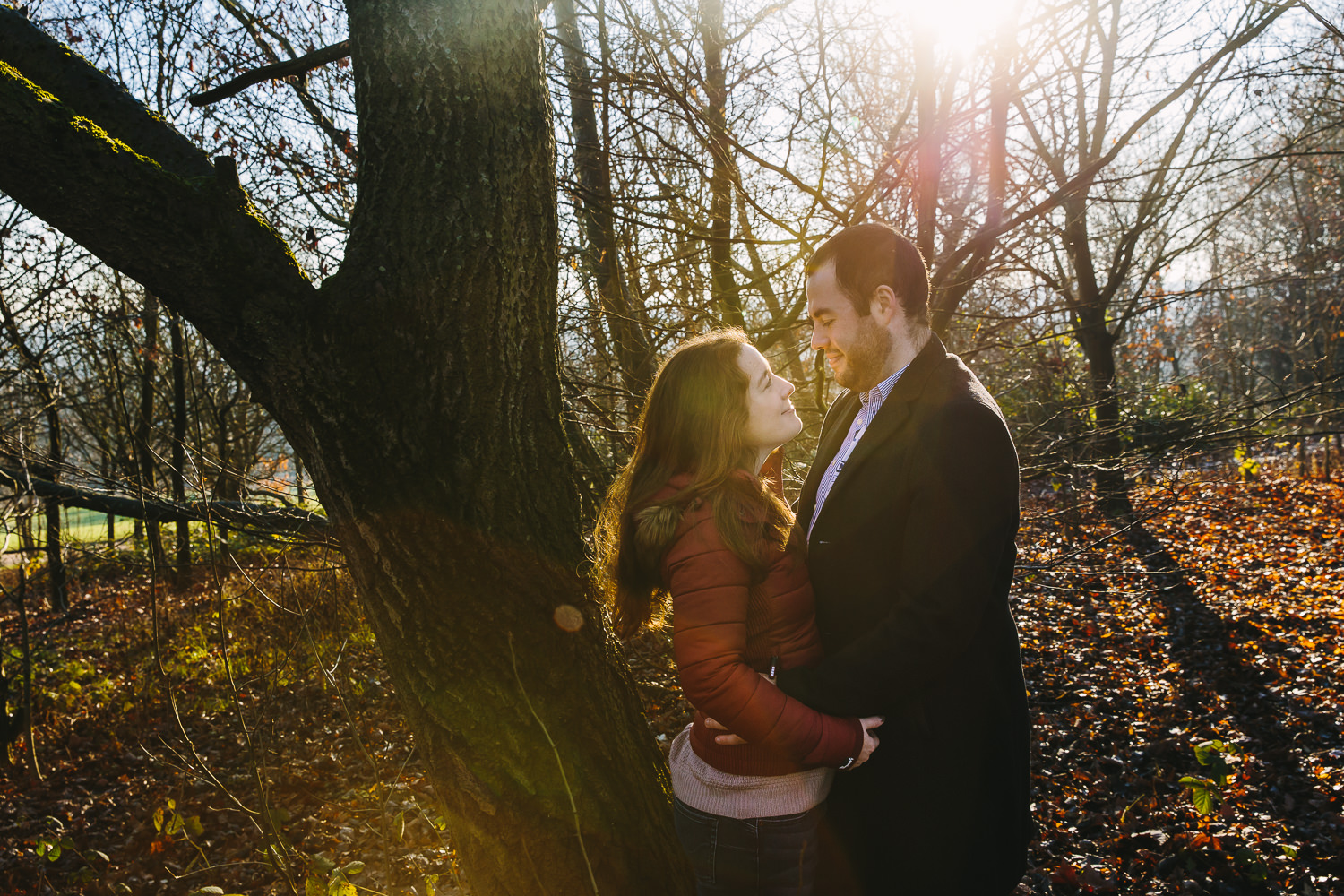 ENgagement shoot photo of couple smiling at each other, with low sunlight shining behind them. They are surround by trees and smiling at one another.