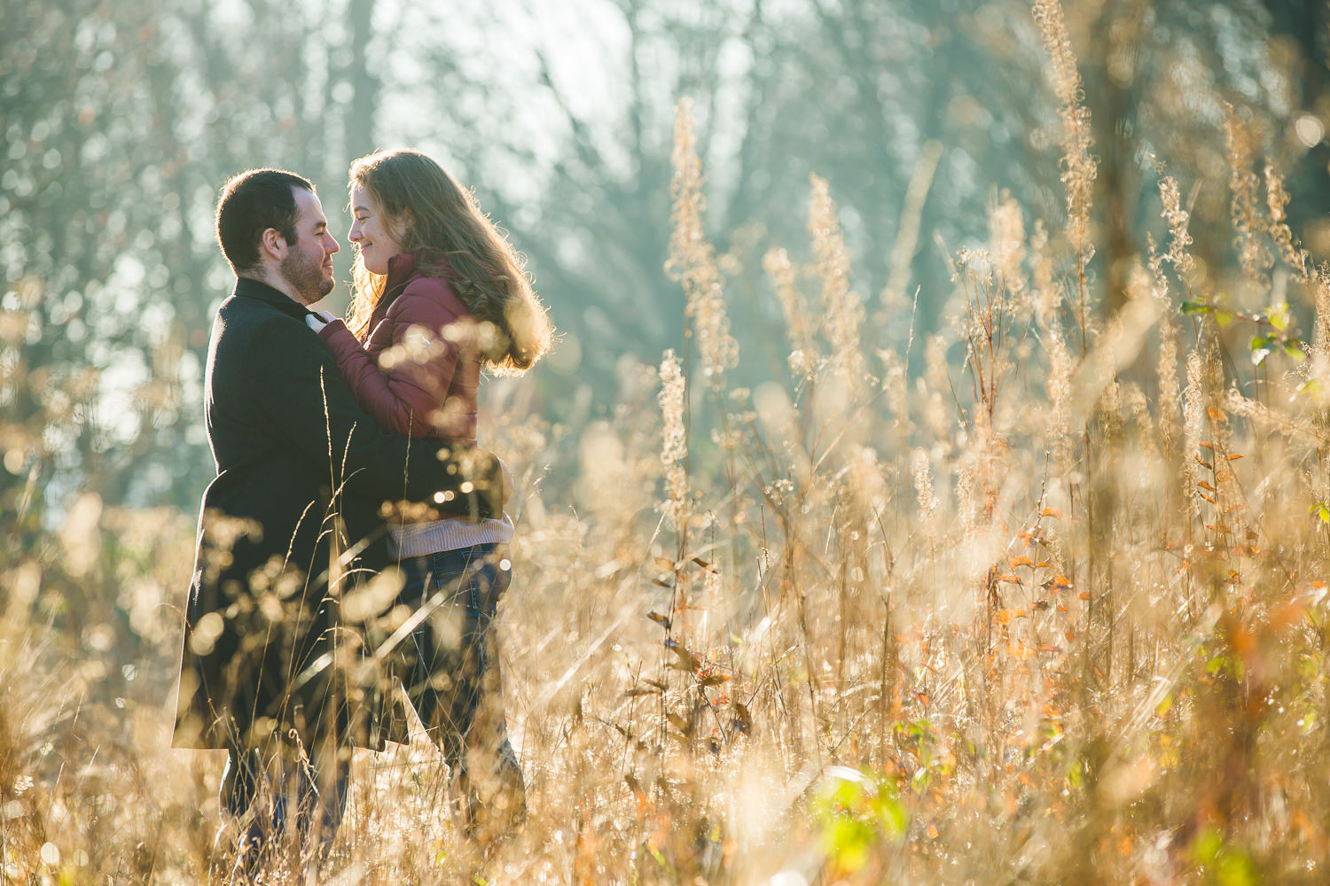 Winter engagement photography of a couple in a field smiling at one another. with low sunlight shining through the trees and foliage