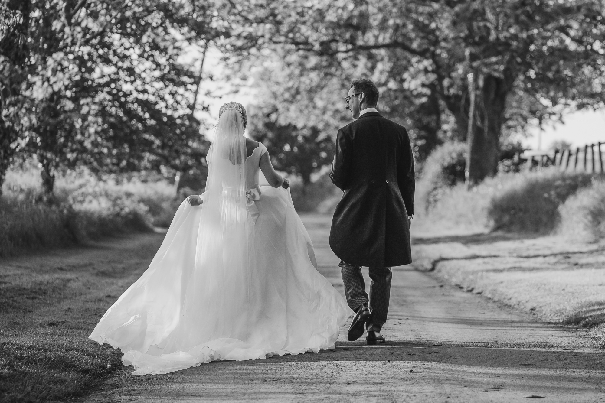 A black and white image of bride and groom walking away from the camera along a country lane.